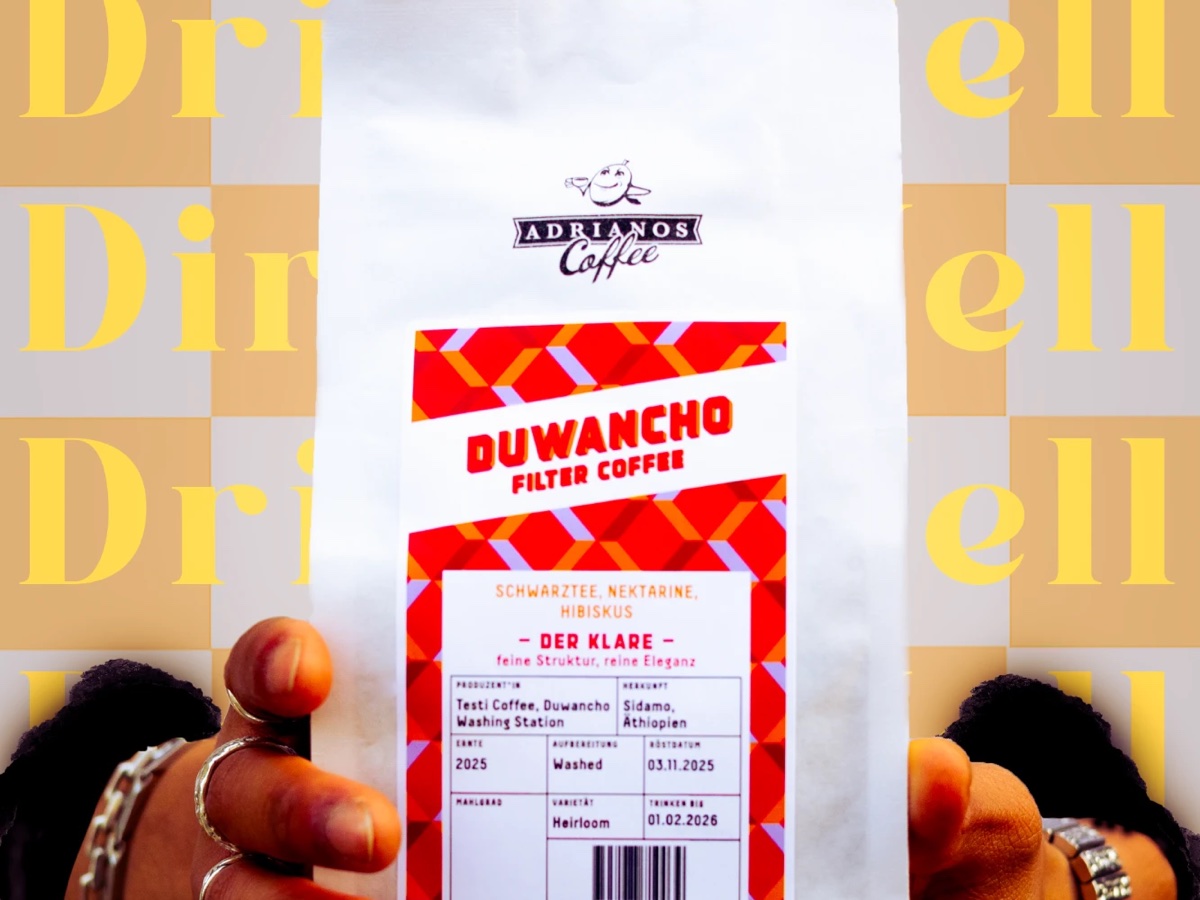 Zwei Hände halten eine weisse Kaffeeverpackung mit rotem Etikett «Duwancho Filter Coffee» vor einen Hintergrund, auf dem «Drink Well» in gelben Serifenbuchstaben wiederholt ist; von oben fallen Kaffeebohnen, halbierte Nektarinen und Hibiskusblüten.